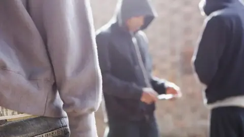 Getty Images Two men in hoodies stand together in the street while a third figure approaches them from the foreground. One of the men is holding something indiscernible in his hands.