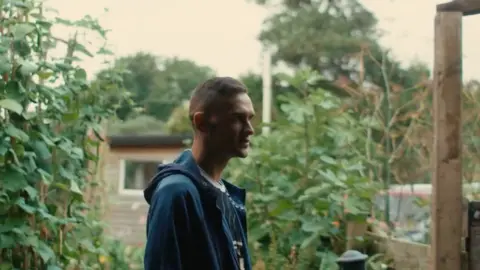 Rob Yates stands in a garden wearing a blue jacket. He has short brown hair and looks to the right of the camera.