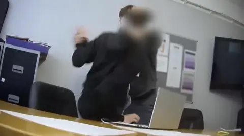 Undercover footage shows a man holding a pupil's hand in the air next to a desk with a laptop and sheets of paper in a classroom