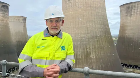 Mike Lewis, CEO of Uniper, stands in front of cooling towers and is wearing a yellow hi-vis jacket, safety glasses and a white hard hat.