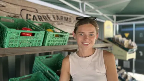 A woman stands beside shelves of fresh vegetables in a covered farm shop area, with green produce crates and a "Carré Farms" sign visible in the background.