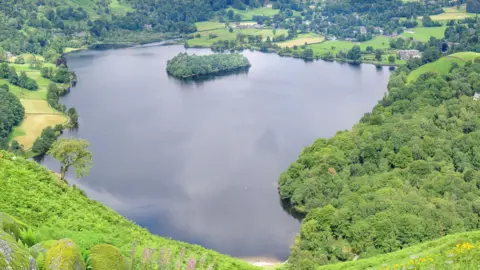 Grasmere, which is a lake with a small island towards the back left. The island is covered in trees and the water on the lake is calm. Bright green fields and trees surround the perimeter of the lake. Houses and buildings are scattered across the landscape.