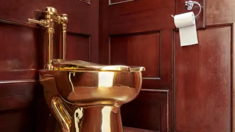 A shiny gold toilet in a dark brown, wood panelled room. A white toilet roll hangs the wrong way on the wall.