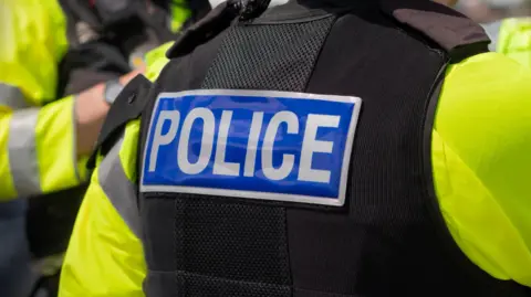 The image shows a close-up of POLICE marking written on the back of a hi-visibility stab proof vest worn, by a trio of police officers at the scene of an incident. 