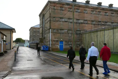 people walk through a yard outside an old prison block