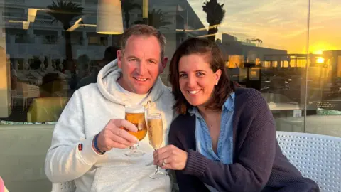 Lucy Lucy and her husband Tom, who is a farmer. They are smiling at the camera and doing a 'cheers' with a beer and a glass of prosecco.