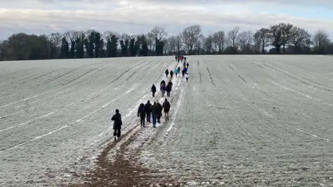 Mr T A line of walkers on a footpath through a frosty field. There is a hedgerow of bushes and trees on the far side of the tree. 