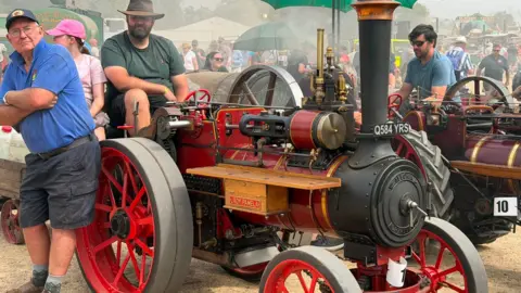 Sally Williams "Restored miniature steam traction engine with bright red wheels and a black chimney on display at a steam rally. Several visitors sit beside and behind the engine, while a large green umbrella provides shade. Steam drifts across the busy exhibition ground in the background."
