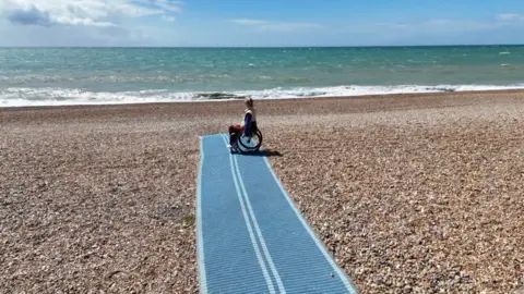 Juliette Parkin/BBC A woman in a wheelchair. She is on a boardwalk at a pebbled beach.