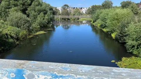 a photo taken from the footbridge looking down onto the empty river with the weir at the end and trees on either side.