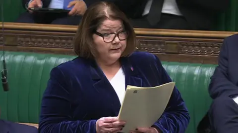 Julie Minns is a woman of about 40 wearing a blue velvet jacket and a white top. She has brown hair and glasses and is reading from a speech in the House of Commons. Behind her you can see the green benches and the legs of the MPs behind her. 