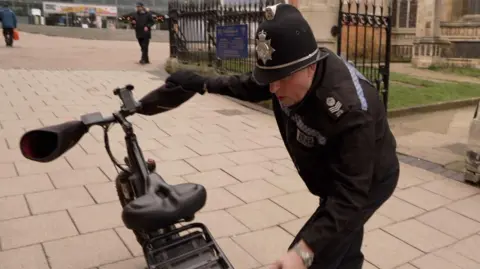 Shaun Whitmore/BBC A police officer is looking at a black bike. He is stood next to the bike and is wearing a black police uniform and hat.