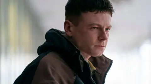 BBC/Mam Tor Productions/Anne Binckebanck Stuart Campbell is in character as Young Ruben. He is straight-faced and stern, watching something in the distance, with a brown and black coat pulled up around his neck.