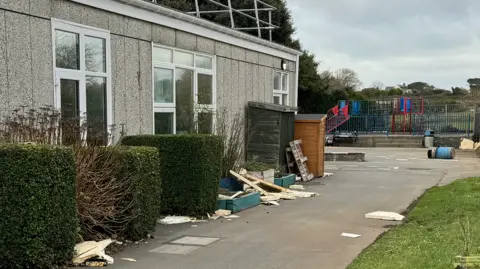 There is debris strewn all over the ground surrounding the school including wood, slabs and roofing material.