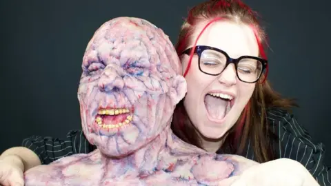 A woman in glasses pretending to scream posing behind a bust of a humanoid monster. The monster has yellow teeth, pink-purple skin and visible veins.