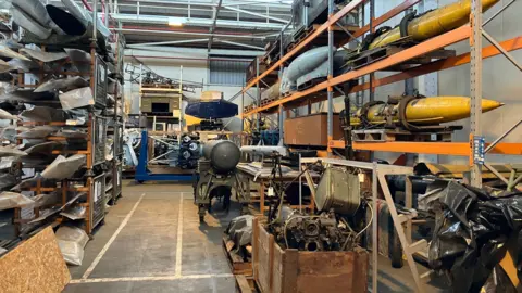 Historical RAF items including bombs, parts of planes and various other equipment in a warehouse. Some are on shelves while others are in boxes.