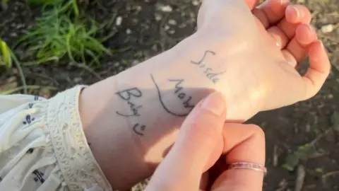 Family Photo An image of Mary's left wrist showing the tattoo of Alex's handwriting. The photo is taken from above, there is grass underneath Mary's wrist in the background and she has a white floral blouse which has been pulled up her arm slightly to show her wrist. Her right thumb is positioned as if pointing towards the word 'Sisda' and she has a pink gemstone and silver band on her right index finger. Another part of the tattoo which is in vision reads 'Bucky' with a smiley face.