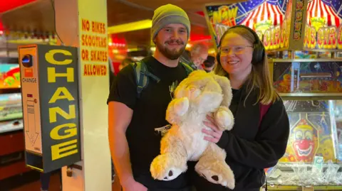 A smiling couple hold up a large bunny they have won at the fair. Coin pusher games are in the background.