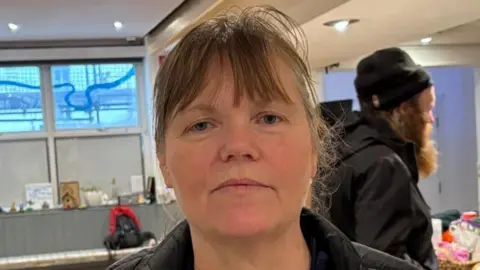 Charlie Stubbs/BBC News There is a woman in a black coat inside of a homeless shelter. She has brown hair and blue eyes. There is a man in the background who is homeless. He is wearing a black coat , a black beanie and a big brown beard. 