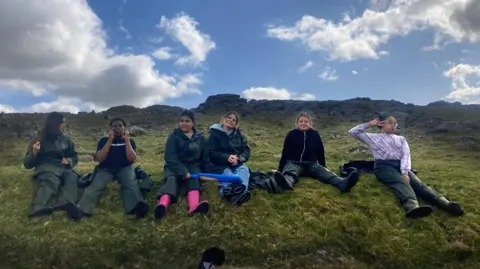 Six girls in wet weather gear sit on the grass on the side of a hill, with granite outcrops behind them