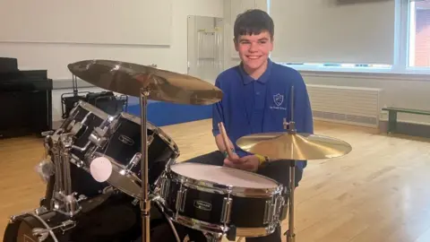 Keelan is wearing a blue polo top and is smiling at the camera. He has short brown hair. He is sitting at a drum kit holding drum sticks.