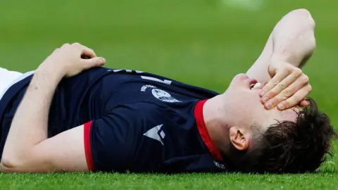 George lies on the pitch and has a hand over his face. He is wearing Ross County's dark blue home kit.