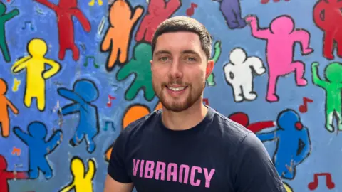 Mr Squire is smiling at the camera while wearing a top with the words 'Vibrancy' written on the centre in pink. He has short brown hair and a beard. Behind him is vibrant blue board with drawings of figures dancing. These have been coloured in blue, red, pink, yellow, orange, white and green.