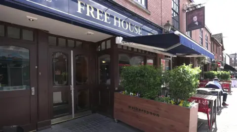 The outside of a pub which has chairs outside and a brown wooden door at the entrance. There is a sign with a man's head on it and planters of flowers, one which says Wetherspoon" on it. 