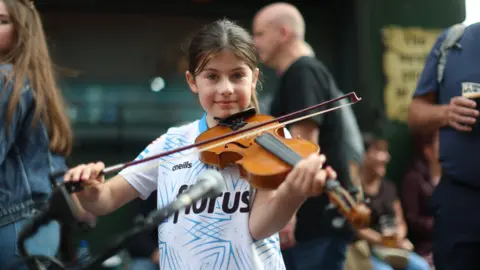 PA Media A girl is playing the fiddle. Her brown hair is in a ponytail. People are in the background. A man is holding a Guinness in a plastic cup. 