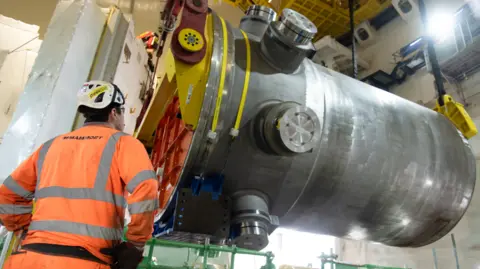 A man wearing an orange high visibility jacket has his arms on his hips, looking at a large metal cylinder.