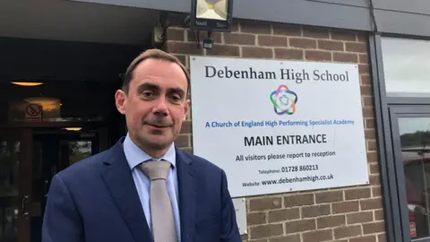 Vikki Irwin/BBC Simon Martin smiles at the camera as he is photographed outside the entrance to Debenham High School. He has short brown hair and is wearing a blue suit. Beside him is a sign indicating it is the main entrance to the school. 