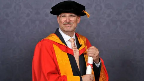 Mark Wheatley holding his certificate and wearing a orange and red university gown. He also has a black cap on. 