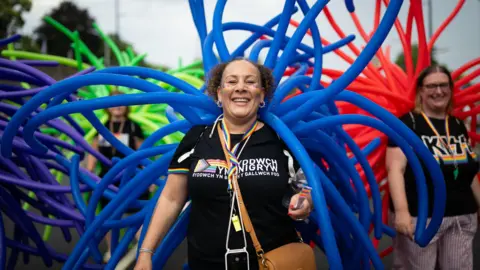 Matthew Horwood/Pride Cymru A woman with dark hair is wearing a black t-shirt with blue tubes attached to her back. Another woman with long blond hair stands behind to the right wearing a black t-shirt with red tubes attached.