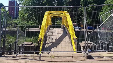 The side of the so-called banana bridge which crosses the Avon New Cut in Bristol. It's a yellow, newly painted bridge, with fences up in front of it before the reopening. 