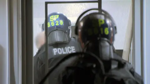 Police officers wearing protective helmets and tactical gear move through a doorway inside a building; “POLICE” and identification markings are visible on their helmets and backs.