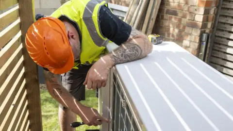 Getty Images A technician in an orange hard hat and yellow high-vis vest leans over a white heat pump unit with a screwdriver in a garden.
