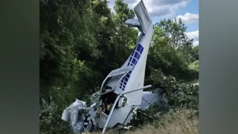 A plane that has crashed into the ground, with trees behind it. There is blue paint on it. The front of the plane is mangled. 