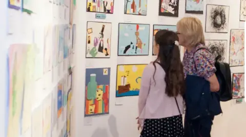 Two white walls are decorated with artwork.  An adult and a child are standing with their backs to the camera, looking at the work.