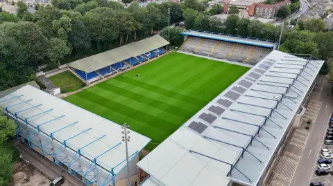 An empty football stadium as seen from above. It has four stands of seat and a green pitch, with a carpark to the right. 