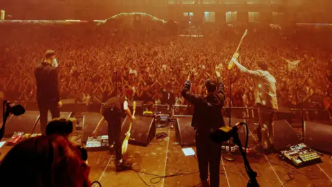Alastair Brookes/KoLAB Studios The band The Maccabees are seen standing on stage in a picture taken from the back of the stage. They are facing a large crowd who are applauding them in the Bristol Harbourside Arena during the Siren festival.
