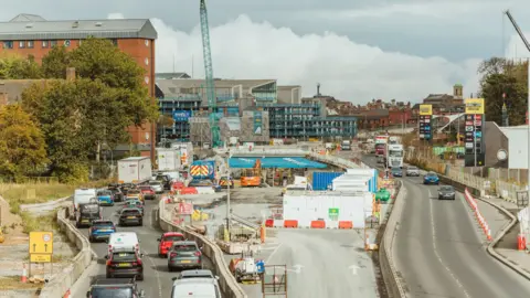 Two roads with two lanes with roadworks going on in the middle. There are multiple cars in the lane on the left. There are buildings and a crane in the distance