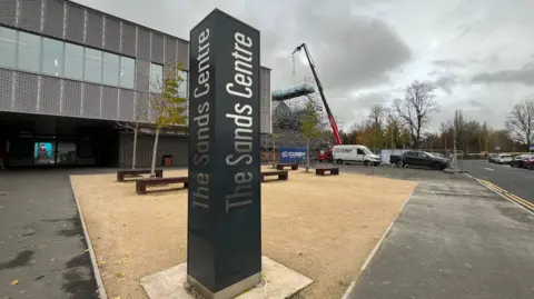BBC/Federica Bedendo A general view of the entrance to The Sands Centre in Carlisle. A black pillar has the name of the venue written on each side in silver. The entrance to the leisure centre is visible on the left, while on the right heavy plant machinery is working on an area of the building surrounded by scaffolding.
