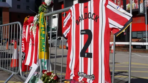 BBC A red and white football shirt with Baldock written on the back, hanging on some grey metal fencing