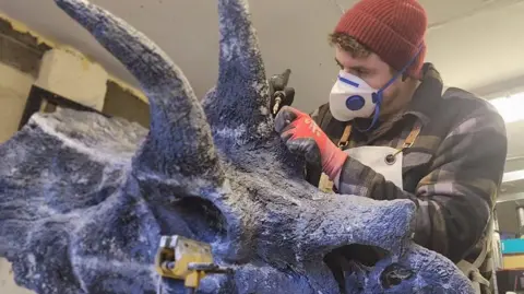 Alex Pritchard Alex Pritchard paints a replica of a dinosaur skull. He wears a mask and gloves as he paints. He wears a red beanie hat and chequered shirt.