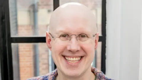 Matt Lucas, wearing glasses, smiles at camera in colourful jumper