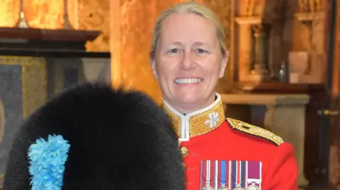 Captain Claire Lawrence, wearing the uniform of the Band of the Irish Guards, and holding a bear skin hat. She is wearing a bright red jacket, with four medals on her lapel.