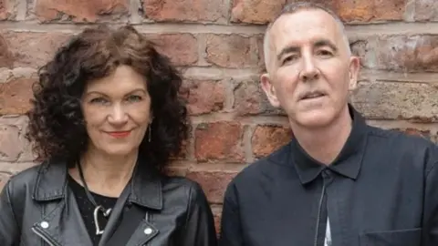 Lilly Shields Alison Bell has dark red hair and is wearing a black motor bike jacket. Liam Walsh has greying hair around the temples and is wearing a dark blue jacket