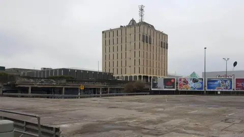 LDRS The former Bonny Street police station: a large, square building behind a large, empty, car park with three big billboards to the right