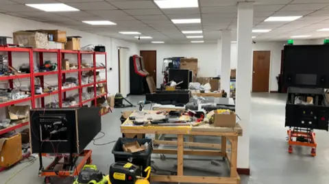 A large space, with red racks of equpiment to the left, a number of wooden frames in the middle, and a large black framed machine to the right. Two traditional arcade machines sit in the corner of the room
