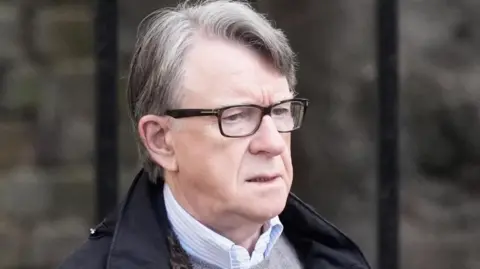 Lord Mandelson wearing glasses, a dark coloured coat, a grey jumper and a shirt.